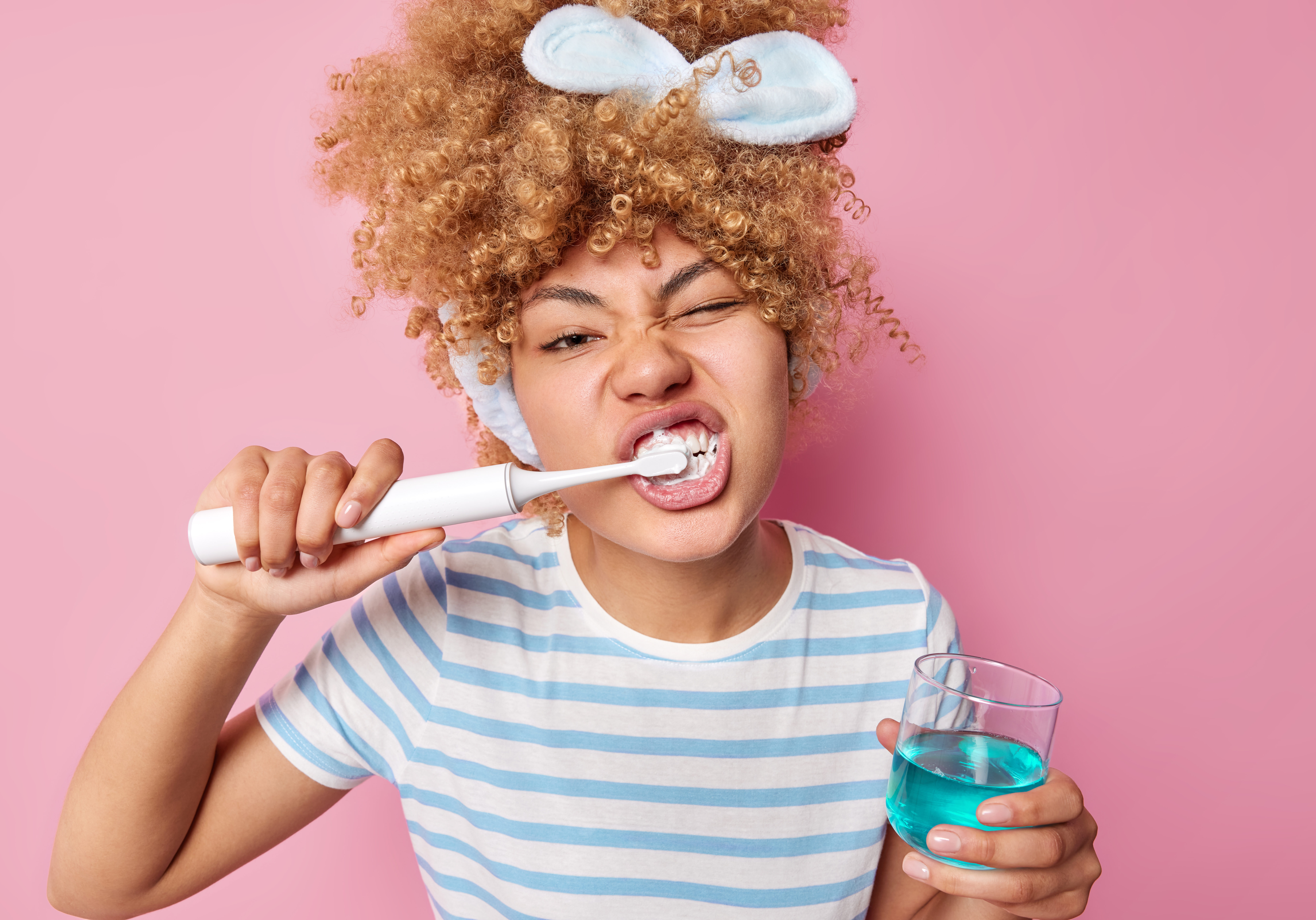 Photo of young woman with curly combed hair brushes teeth uses electic brush undergoes whitening procedure holds glass of mouthwash wears casual striped t shirt isolated over pink background